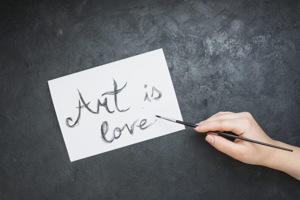 woman-s-hand-written-art-is-love-text-white-paper-with-paintbrush-slate-backdrop
